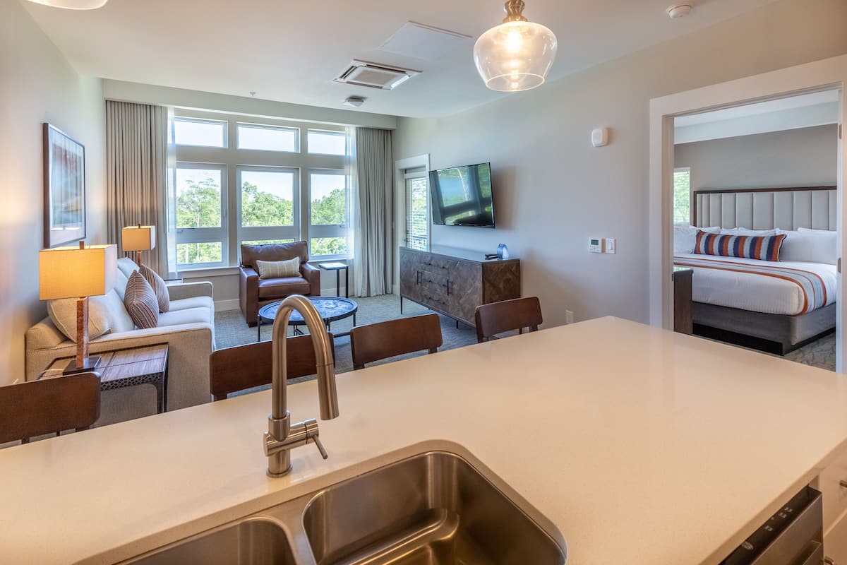 A modern hotel room with a kitchen area, a seating area with a sofa and chairs, a TV mounted on the wall, and a large window offering a view of the outdoors.