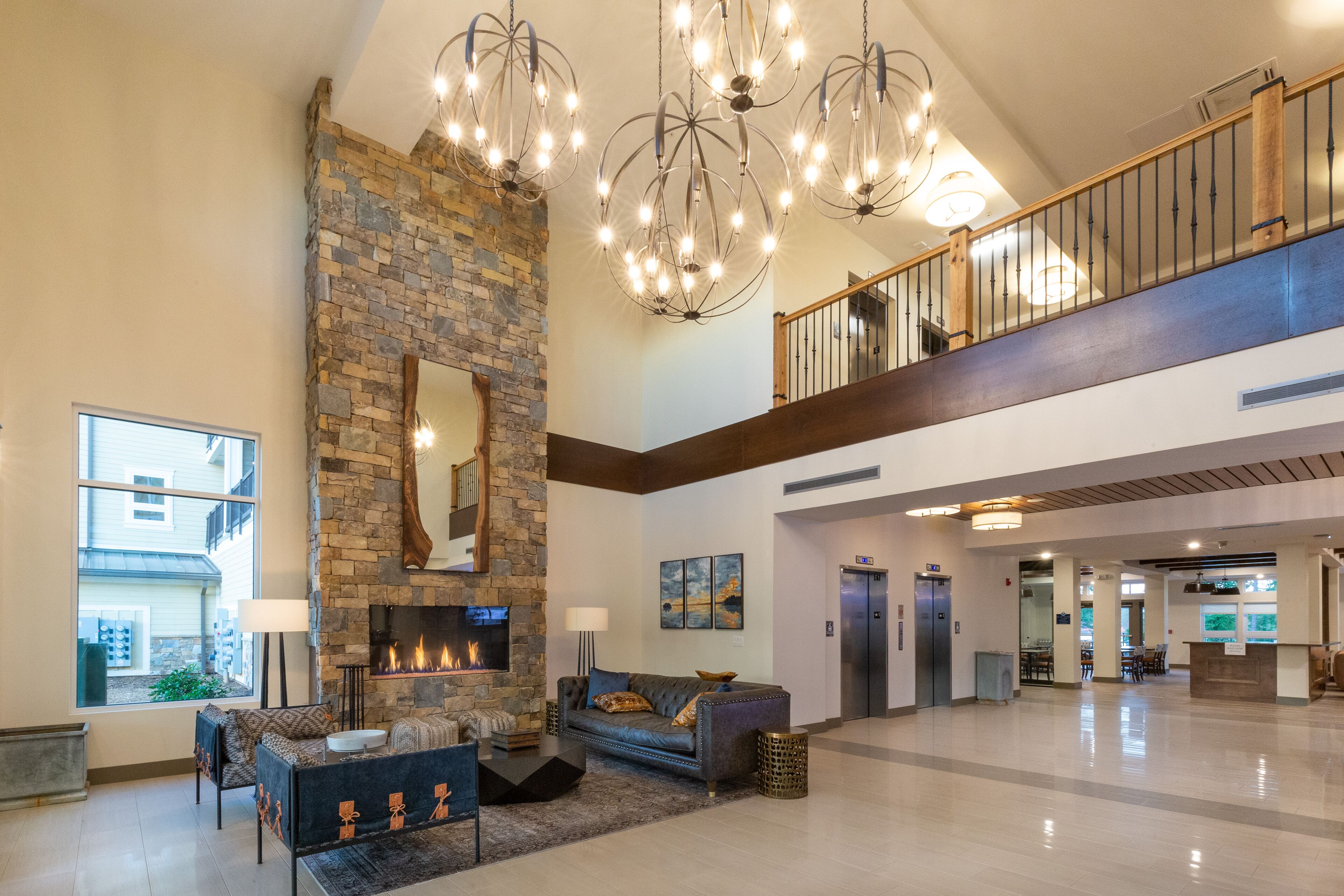 A spacious lobby with a stone fireplace, a high ceiling with chandeliers, a staircase, and a reception desk.