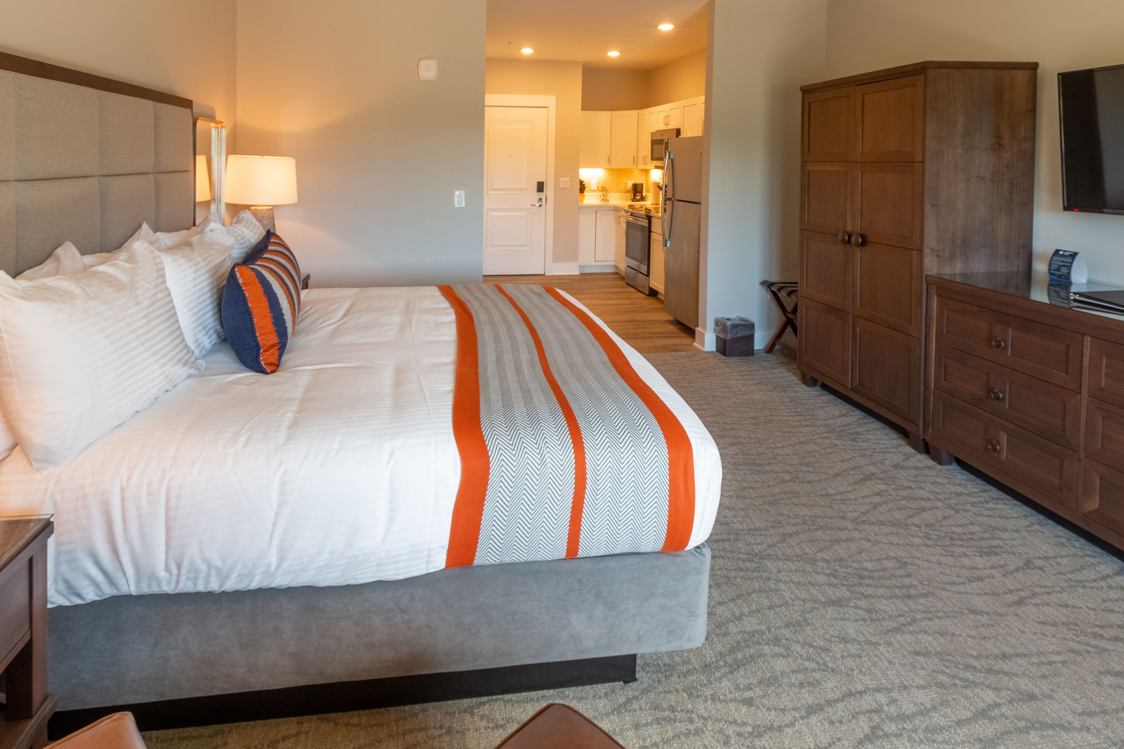 A hotel room with a large bed, a wooden wardrobe, a small kitchen area, and a television on the wall.