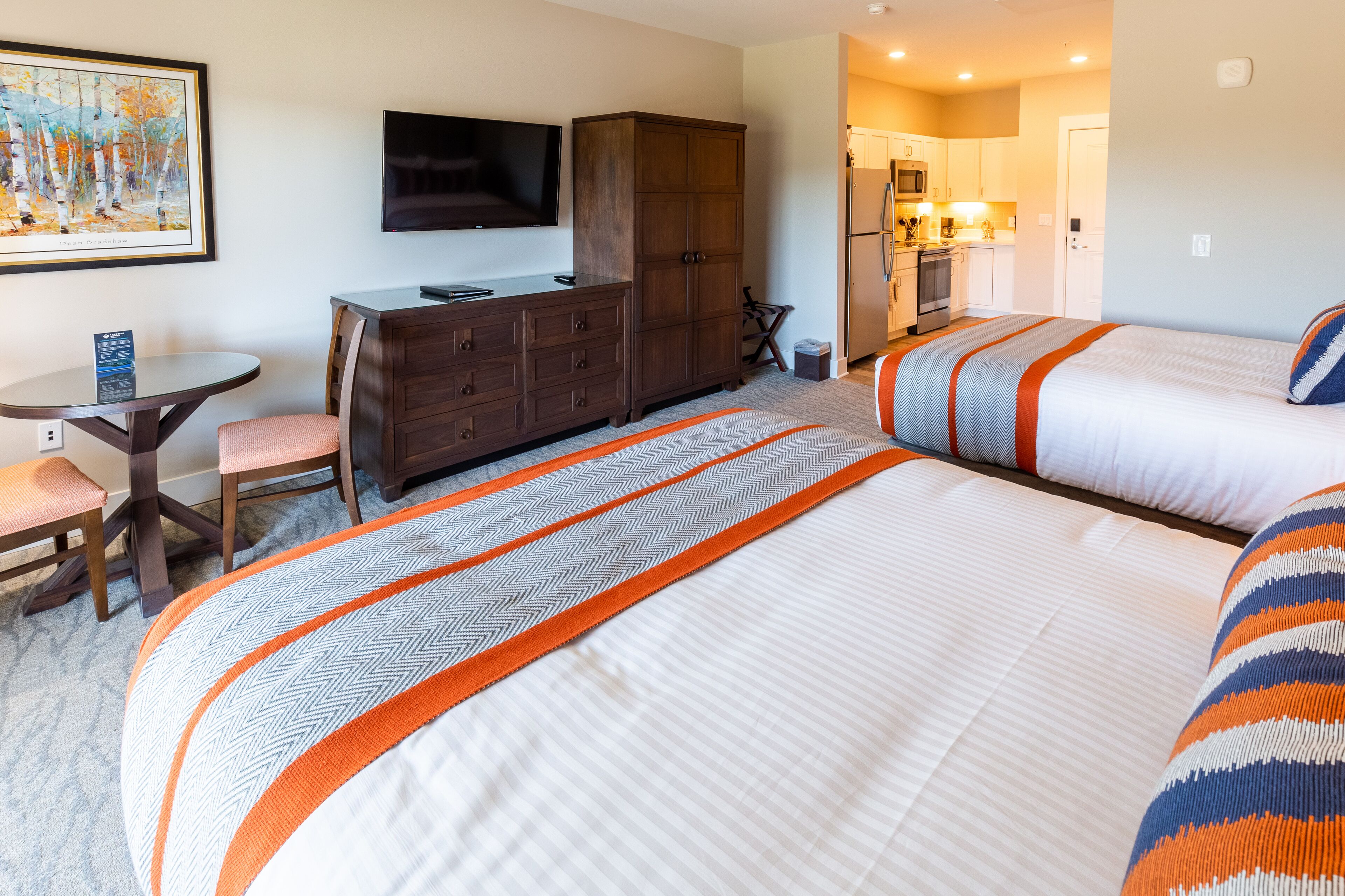 A hotel room with two beds, a desk, a chair, a television, and a kitchenette.