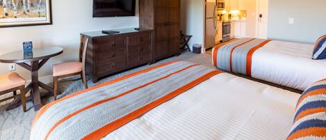 A hotel room with two beds, a desk, a chair, a television, and a kitchenette.