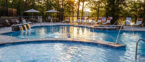 A swimming pool with multiple water features, surrounded by lounge chairs and umbrellas, set in a wooded area.