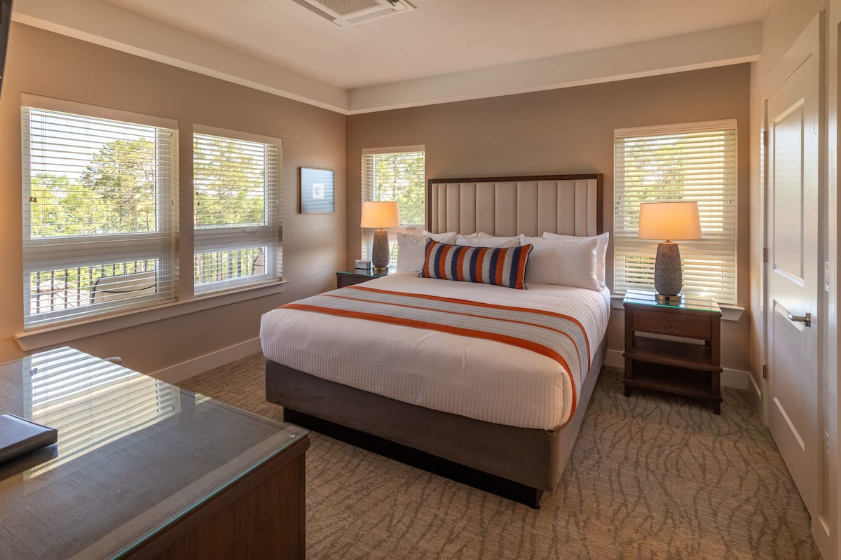 A bedroom with a large bed, two bedside tables with lamps, and windows with blinds.