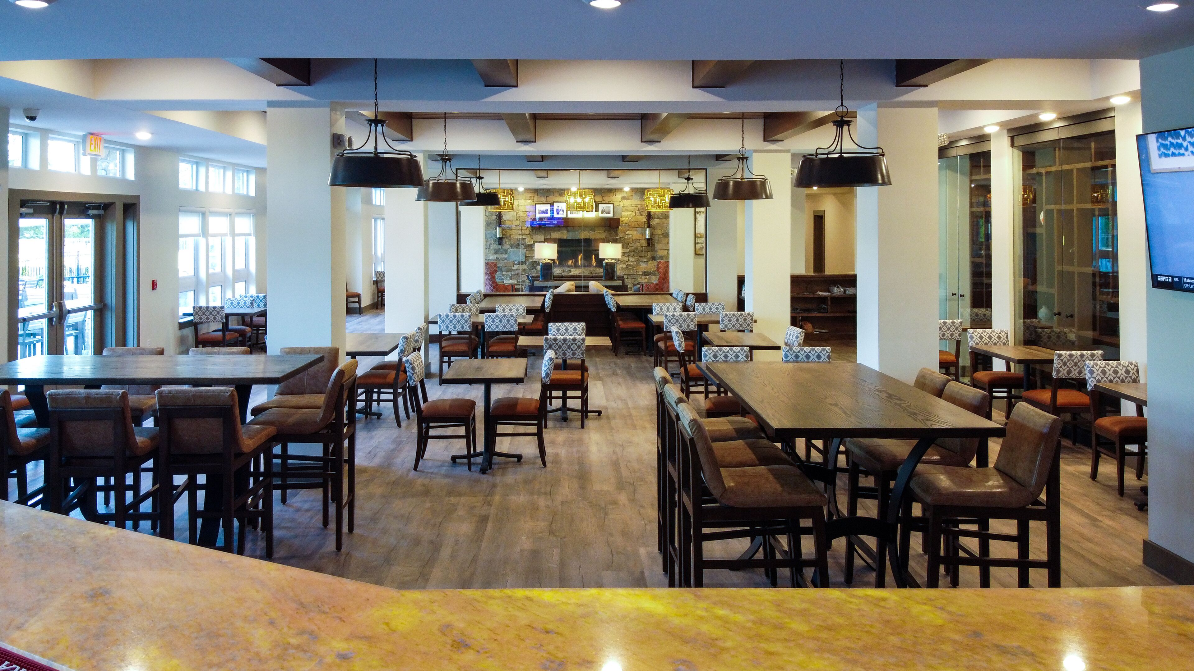 A spacious restaurant with high ceilings, pendant lighting, and a central fireplace. Tables and chairs are arranged for dining.