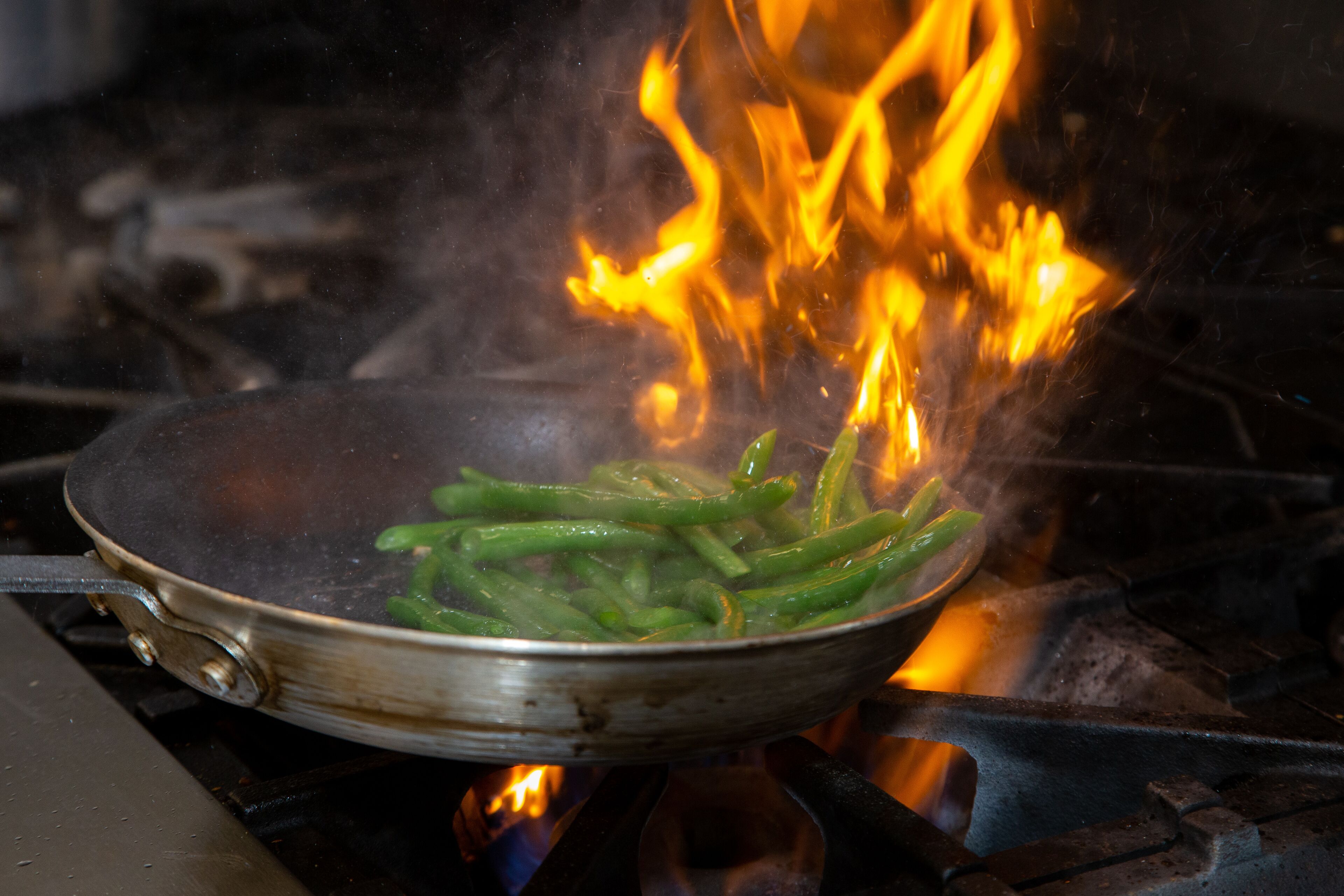 Una sartén sobre la estufa con vegetales verdes y llamas.