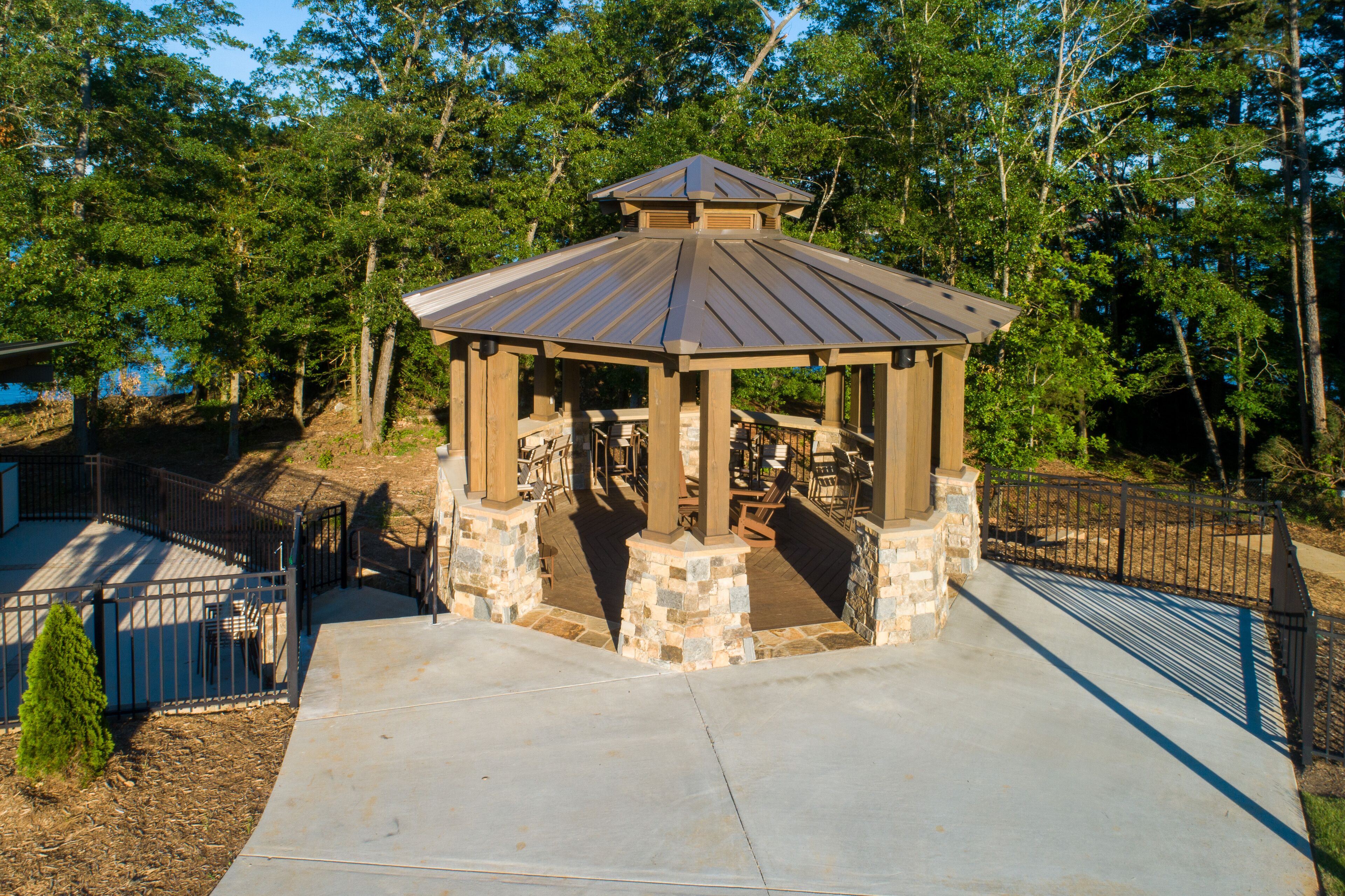 A gazebo with a stone base and metal roof, surrounded by trees and a fenced area.