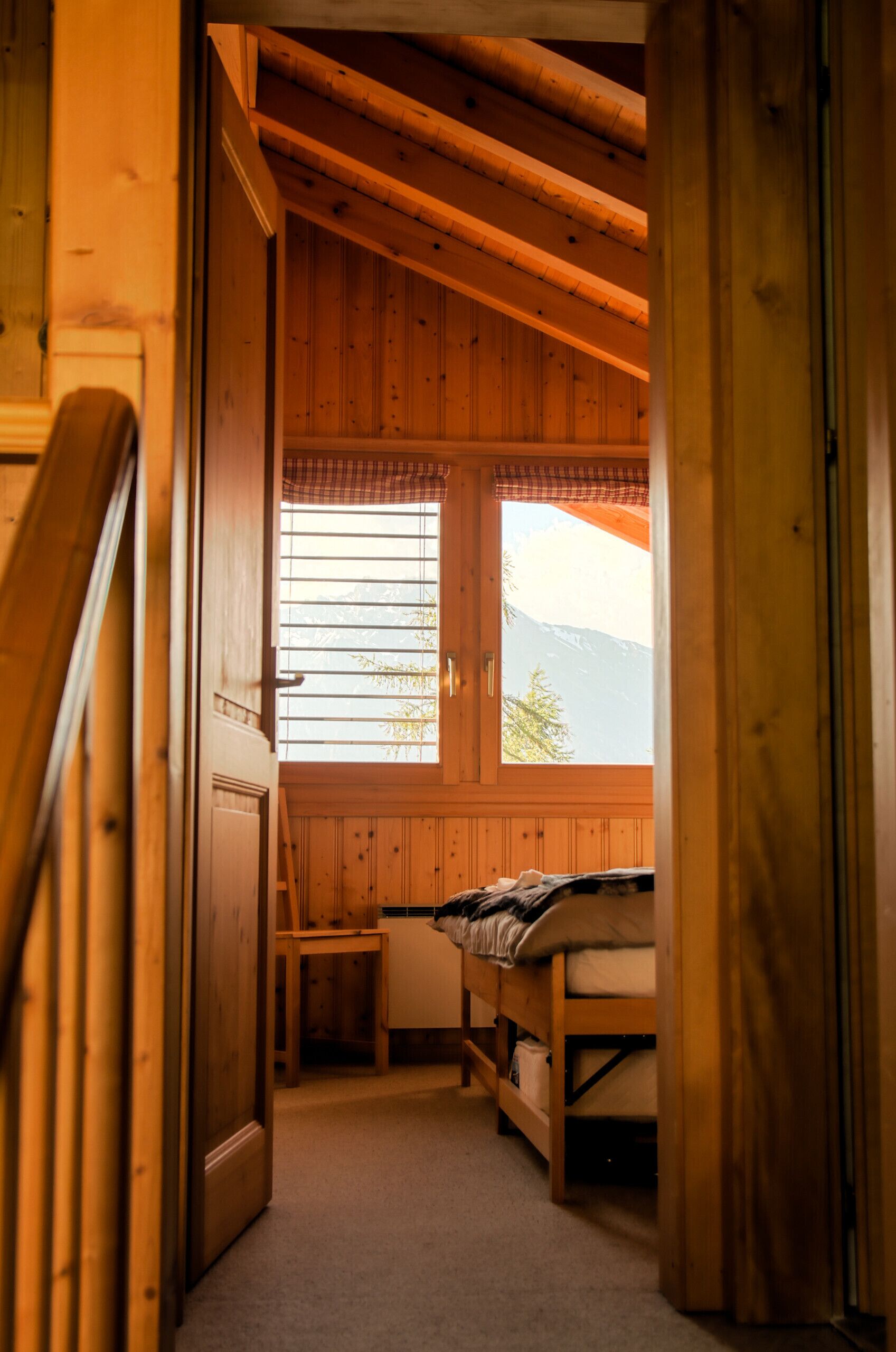A wooden cabin room with a bed, a small table, and a window with a view of mountains.