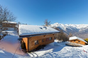 A wooden cabin with a snow-covered roof, surrounded by snow and mountains in the background.