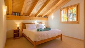 Een slaapkamer met een houten bed, nachtkastje, een plank met boeken, een raam met uitzicht op bomen en een schuine kap.