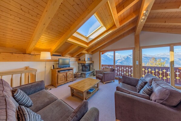 A cozy living room with a fireplace, wooden furniture, and a large window overlooking a mountainous landscape.