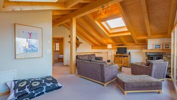 Een gezellige woonkamer met een hoge plafond, houten balken, een bank, een tweepersoonsbank, een televisie en een open haard.
