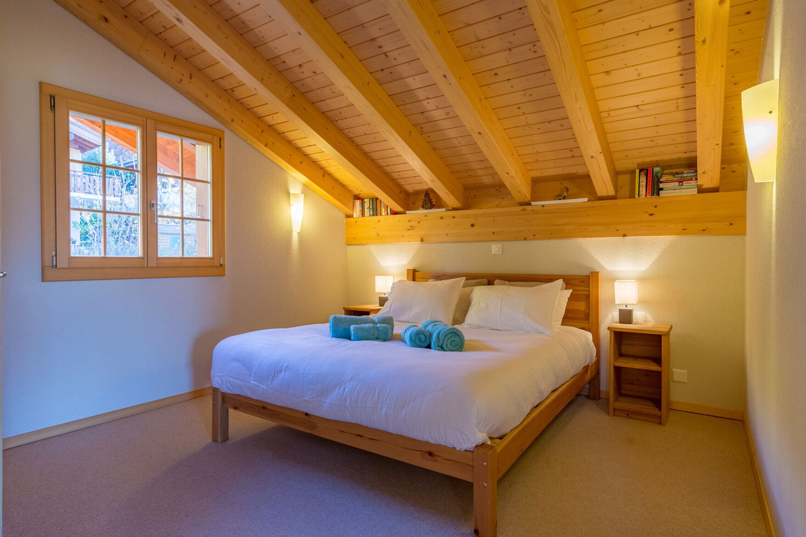 Ein Schlafzimmer mit Holzbalkendecke, einem großen Fenster, einem Bett mit weißer Bettwäsche und zwei Nachttischen mit Lampen.