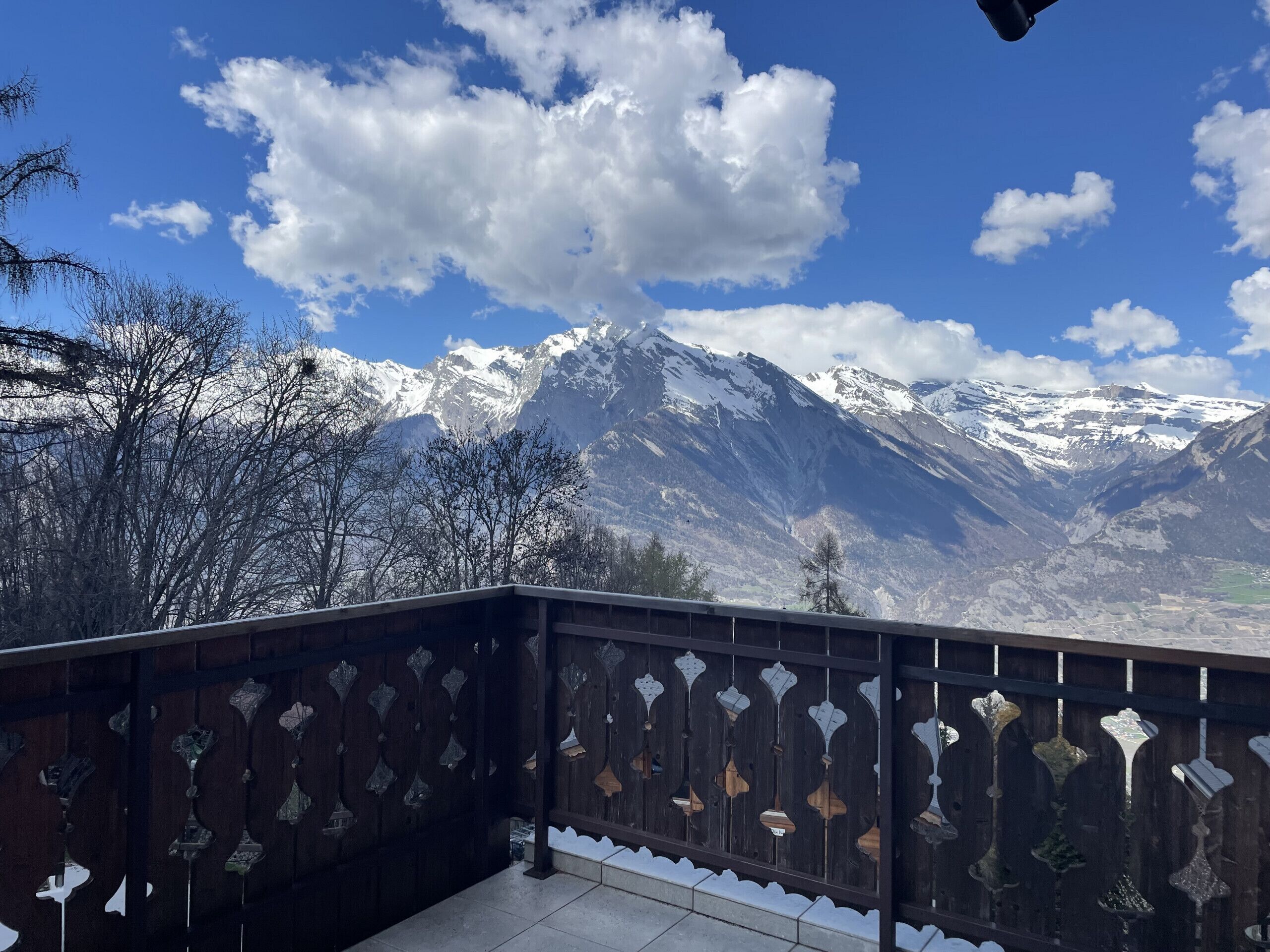Een balkon met houten leuning en hangende decoraties, met uitzicht op een bergachtig landschap met besneeuwde toppen en een helderblauwe lucht.