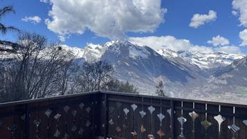 Een balkon met houten leuning en hangende decoraties, met uitzicht op een bergachtig landschap met besneeuwde toppen en een helderblauwe lucht.