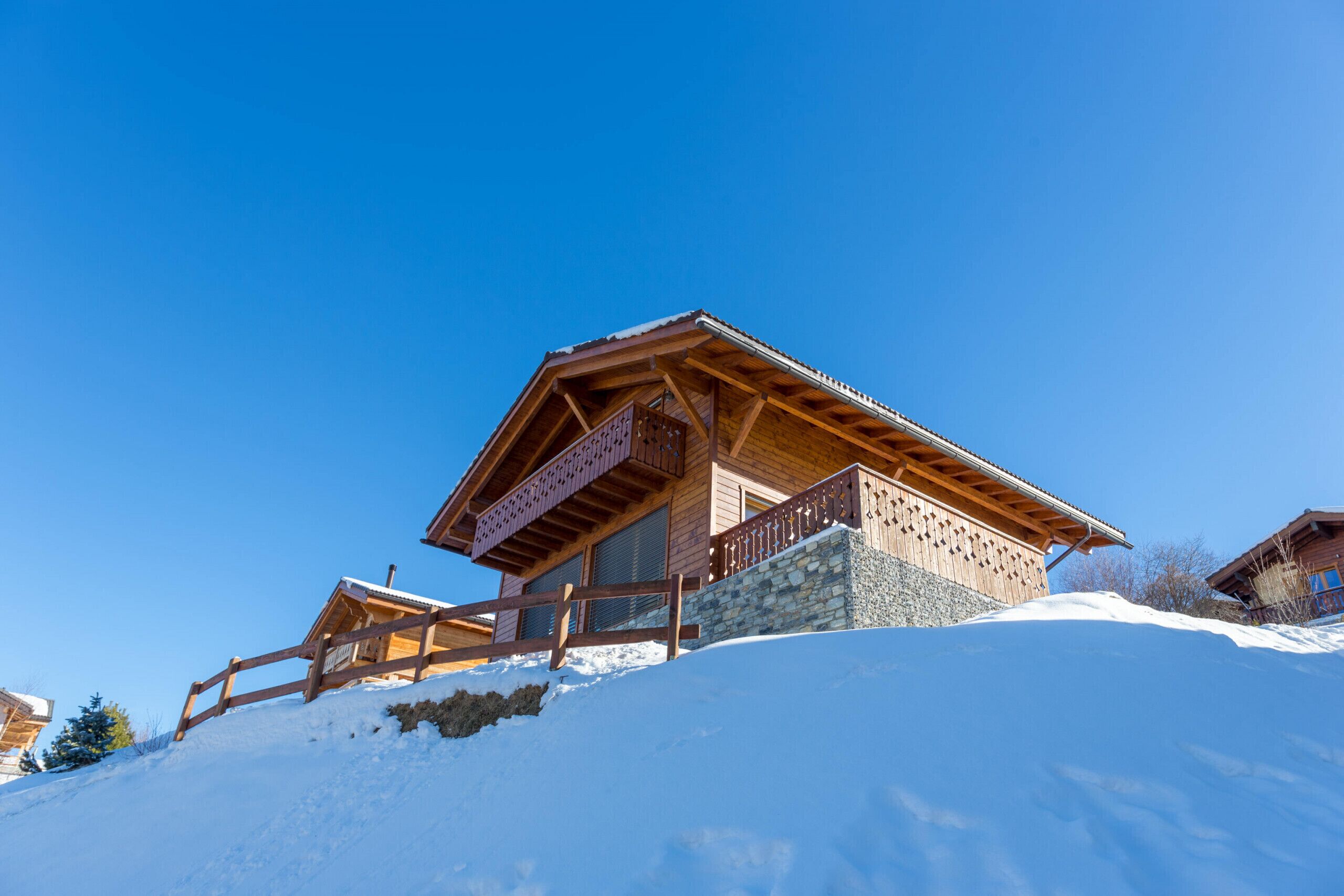 Eine Holzhütte mit Balkon, eingebettet in einen verschneiten Hang unter blauem Himmel.