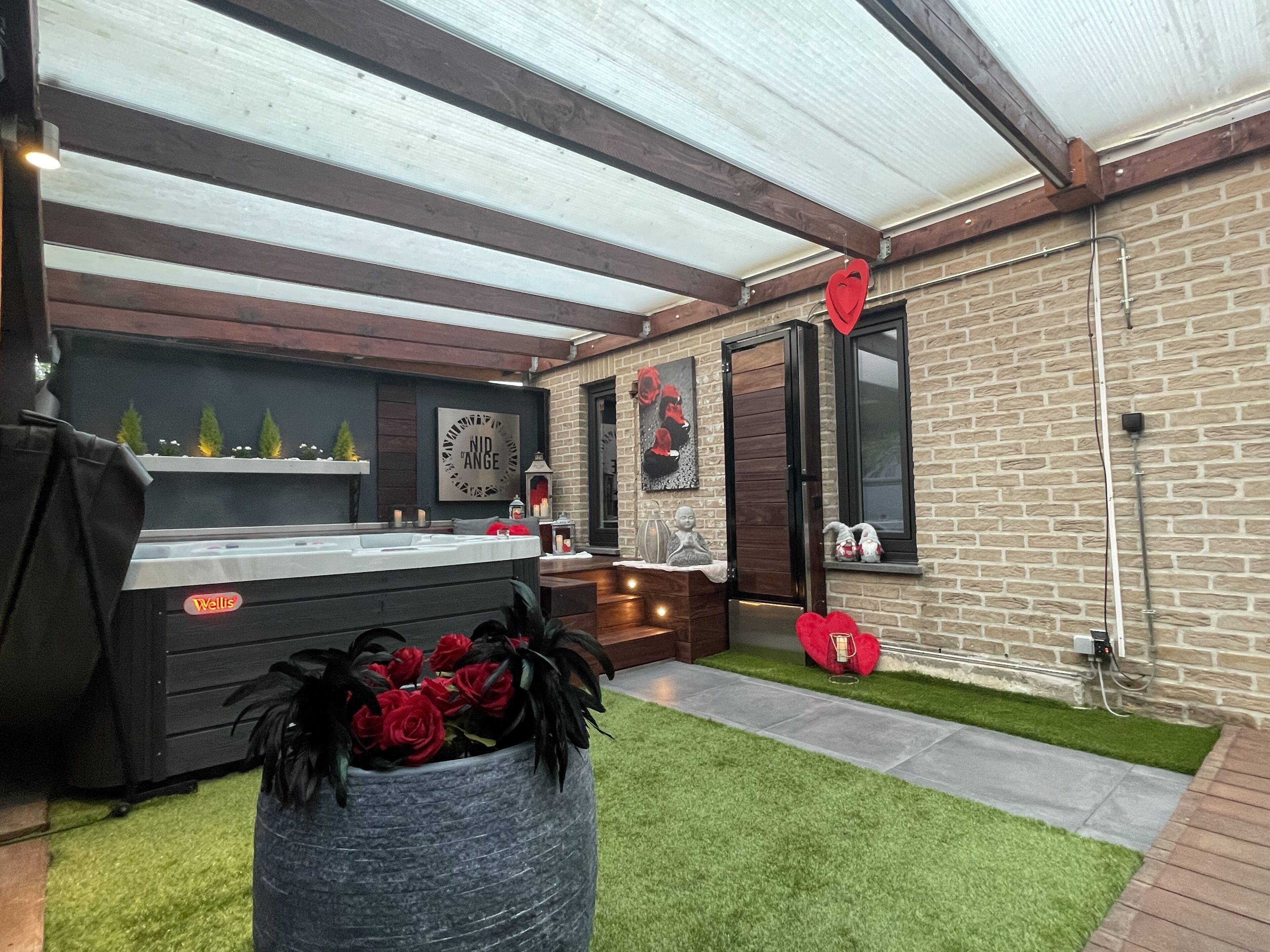 Un espace cuisine extérieur moderne, doté d’un barbecue, d’un jacuzzi et d’un mur décoratif orné d’une enseigne « Nid Angé ».