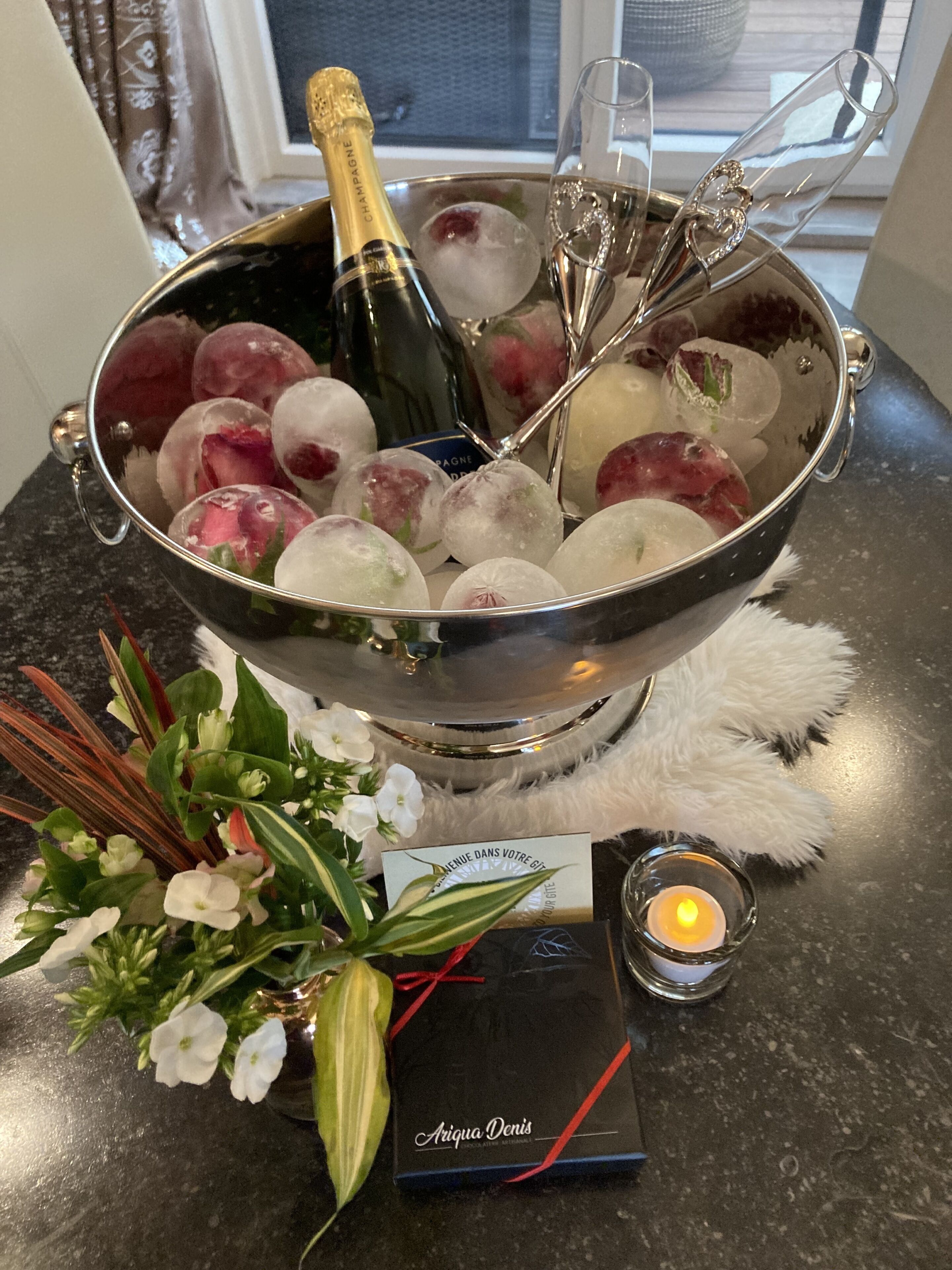 Een champagnefles in een metalen emmer met ijs en een glas, omringd door bloemen en een kaars op een tafel.