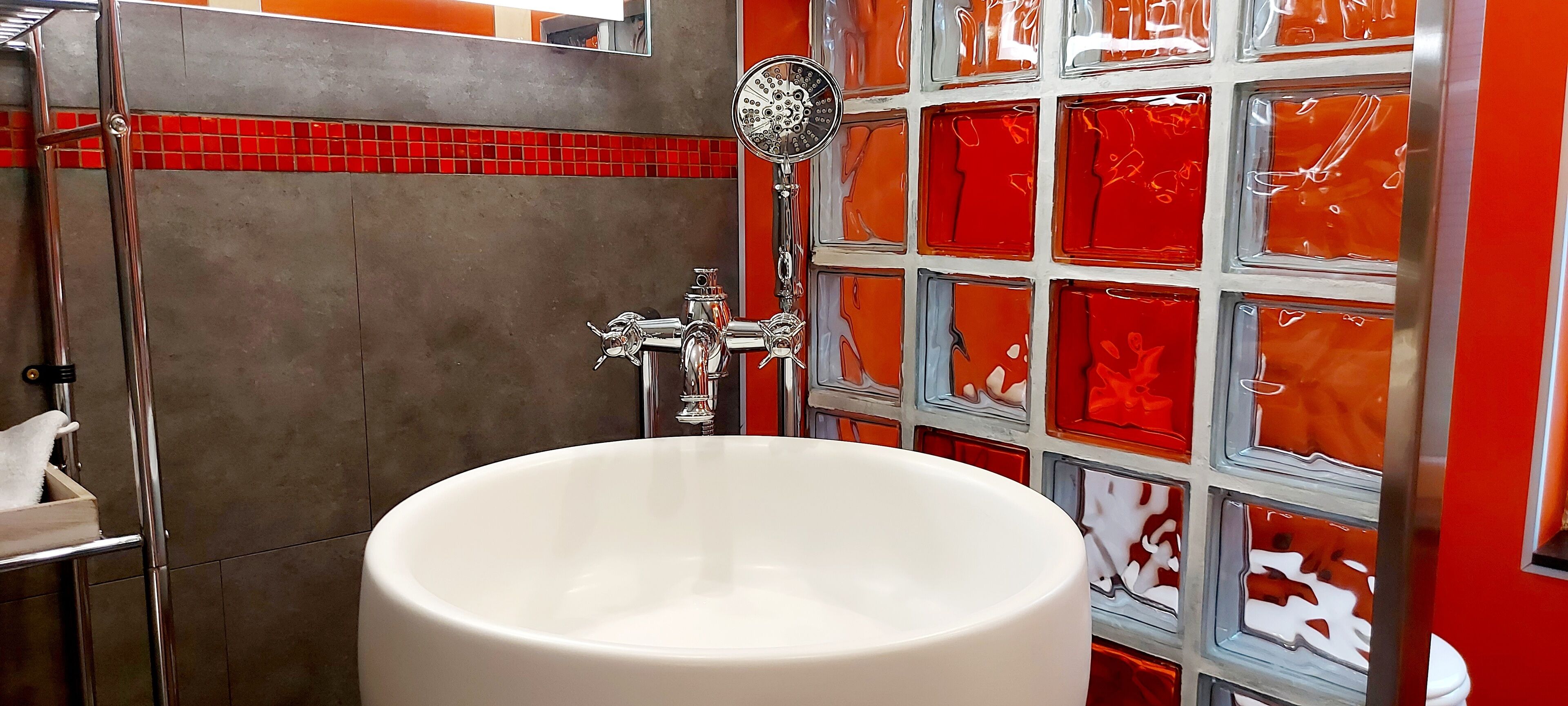 Un baño con una bañera blanca grande, ducha con cabezal de mano y pared de azulejos con diseños rojo y blanco.