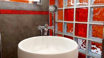 Un baño con una bañera blanca grande, ducha con cabezal de mano y pared de azulejos con diseños rojo y blanco.