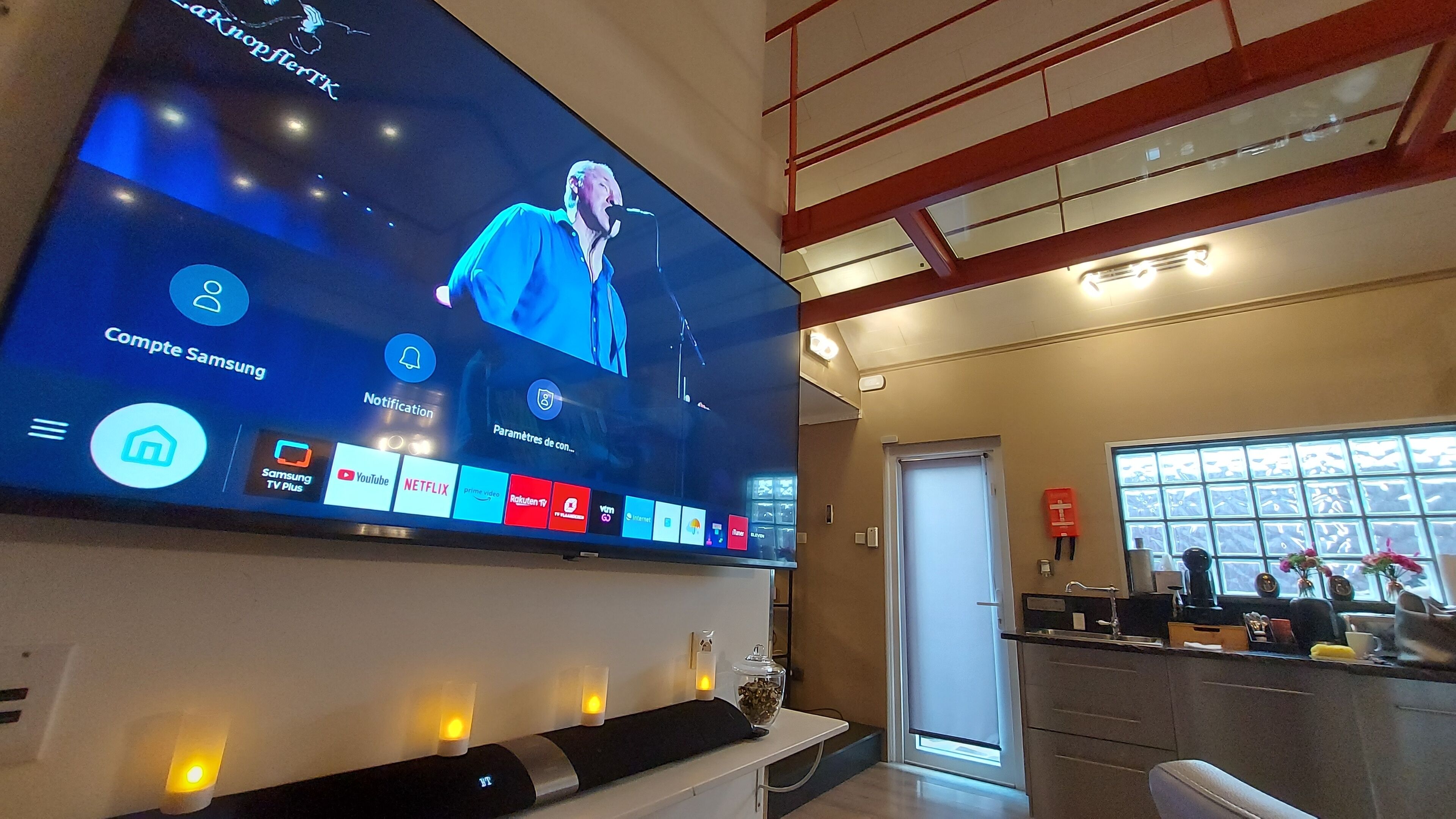 Un grand téléviseur à écran plat est fixé au mur et diffuse un concert en direct. En dessous du téléviseur, on trouve une console blanche avec un plateau noir et un récipient en verre.