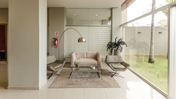 A modern living room with a beige sofa, two armchairs, a glass coffee table, and a large window offering a view of a tree and grass. - Quantum Park (Asunción)