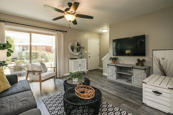 A living room with a sofa, two round black wicker tables, a ceiling fan, a TV, and a sliding glass door leading to an outdoor area. - Updated Condo in A+ Old Town Scottsdale Location! (Scottsdale)