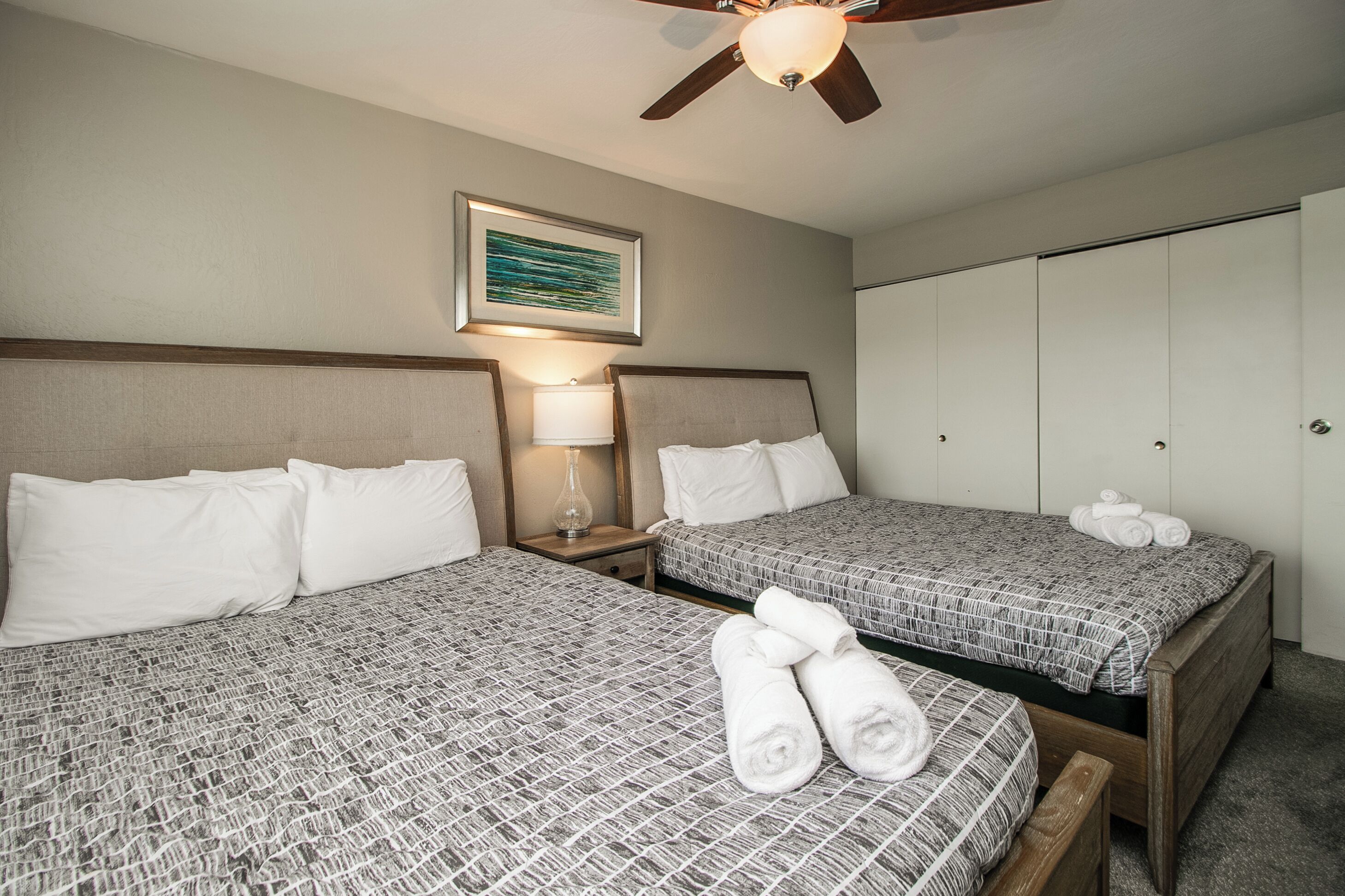 A hotel room with two beds, a ceiling fan, a nightstand with a lamp, and a wardrobe.