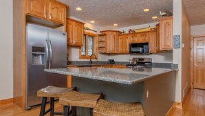 A kitchen with wooden cabinets, a stainless steel refrigerator, a granite countertop, and two bar stools. - High Country Home (McHenry)