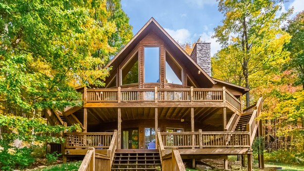 A wooden cabin with a central balcony, a stone chimney, and a wooden deck leading to the entrance. - High Country Home (McHenry)