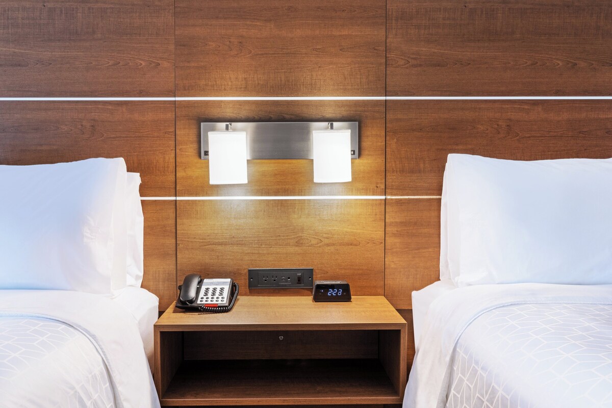 A hotel room with two beds, a bedside table with a phone and a digital clock, and a wall-mounted light fixture.