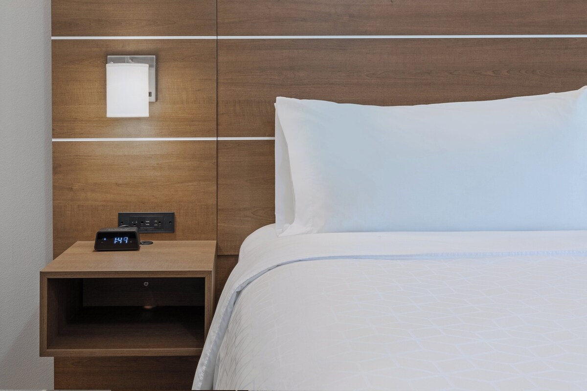 A hotel room with a wooden headboard, a white bed, a nightstand with a digital clock, and a wall-mounted light.