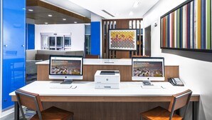 Un espace de bureau moderne avec deux bureaux, chacun équipé d’un ordinateur, d’une imprimante et d’un téléphone. Un tableau coloré orne le mur, et une porte bleue se trouve au fond.