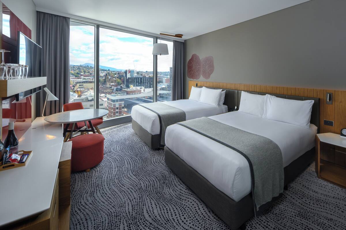 Hotelkamer met twee bedden, een bureau, een stoel, een televisie en een groot raam met uitzicht op de stad.