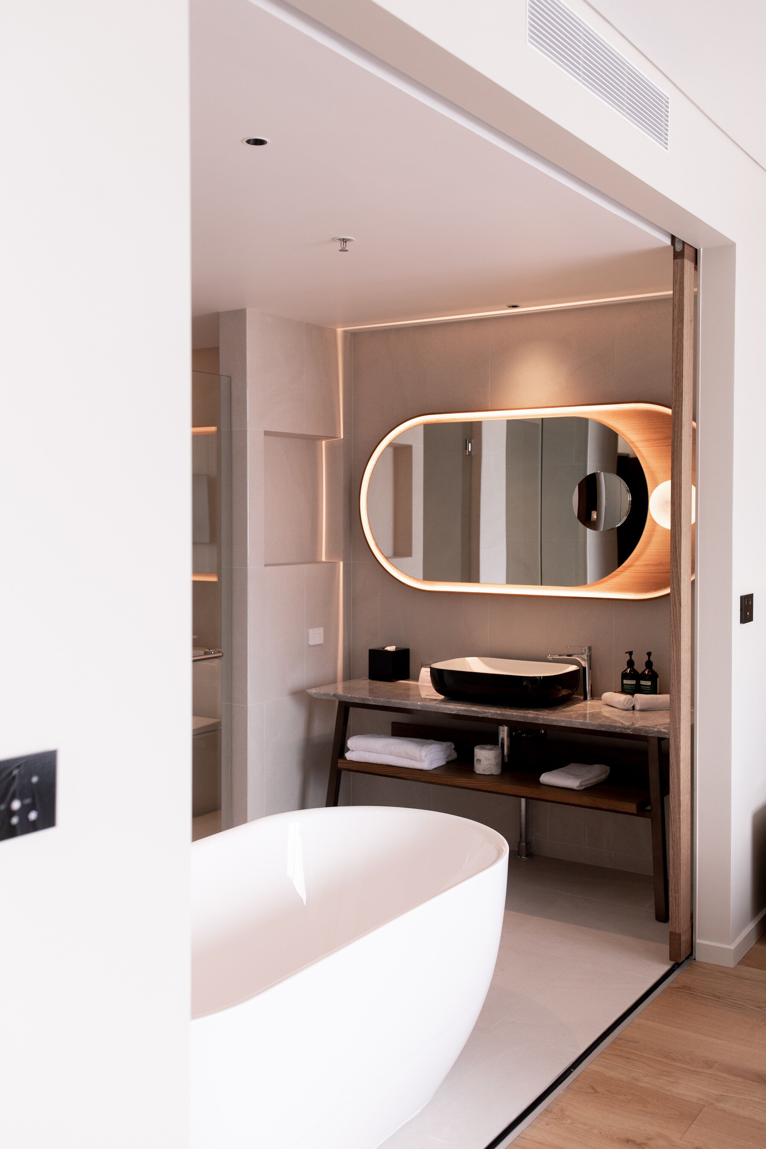 A modern bathroom with a large oval mirror, a freestanding bathtub, and a vanity with a sink and towel rack.