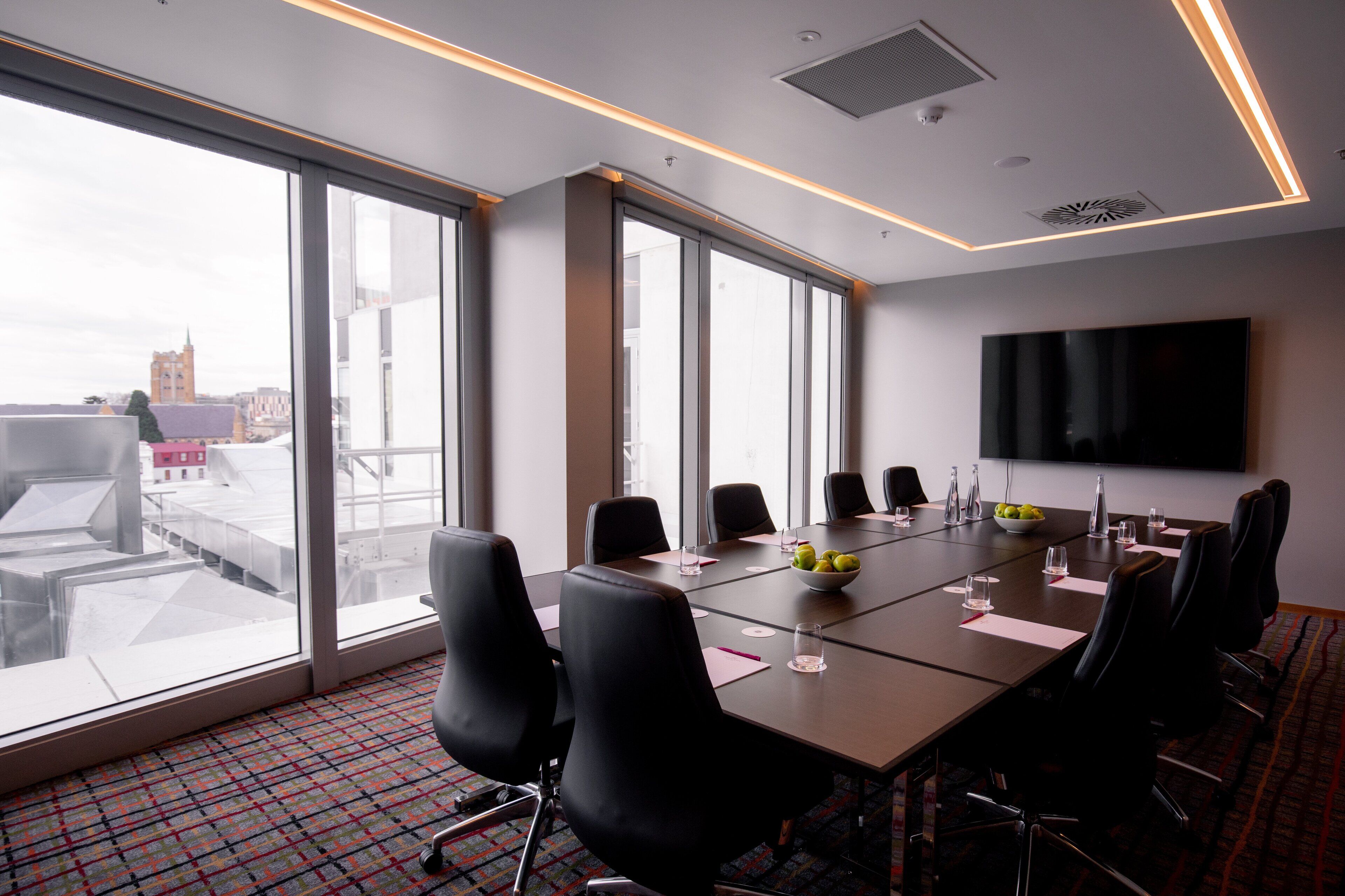 Un salón de conferencias con una gran mesa, sillas negras, un televisor de pantalla plana y ventanas amplias que dan a una vista de la ciudad.