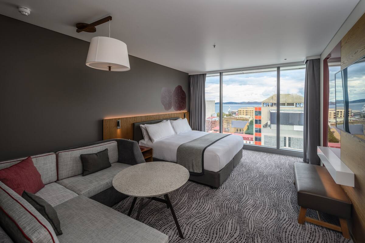 Une chambre d’hôtel moderne avec un grand lit, un canapé, une petite table ronde et une vue sur la ville grâce à de grandes fenêtres.