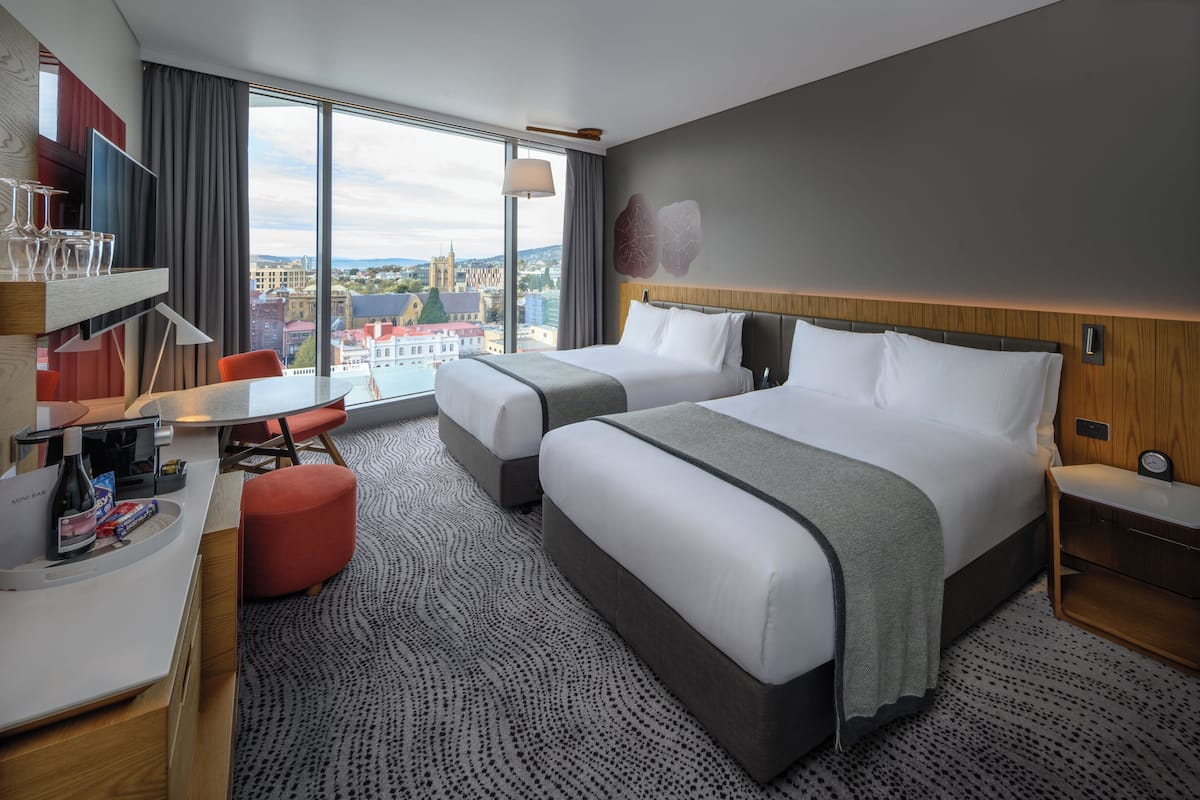Een hotelkamer met twee bedden, een bureau, een stoel en een groot raam met uitzicht op de stad.