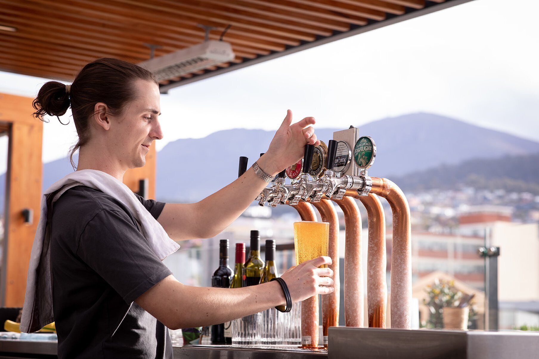 Un bartender sirviendo una bebida de una grifería con varias opciones de cerveza, botellas de vino y una vista panorámica de montañas al fondo.