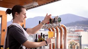 Un bartender sirviendo una bebida de una grifería con varias opciones de cerveza, botellas de vino y una vista panorámica de montañas al fondo.