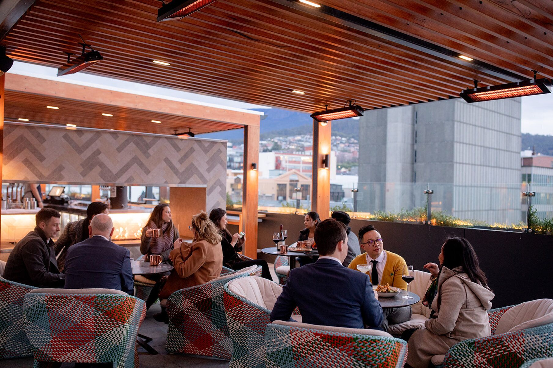 An outdoor seating area with wicker chairs and tables, a bar in the background, and a view of buildings through large windows.
