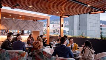 An outdoor seating area with wicker chairs and tables, a bar in the background, and a view of buildings through large windows.