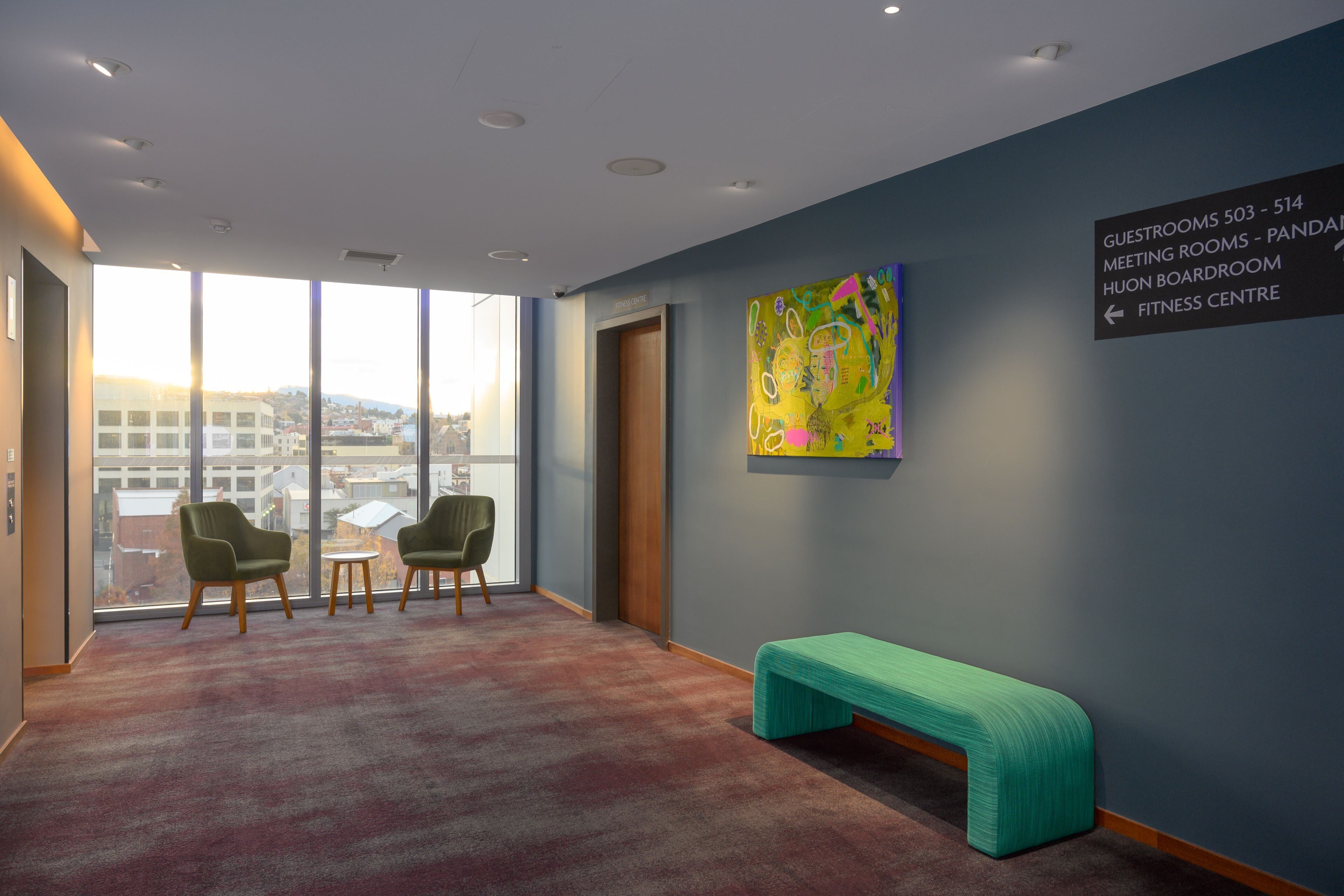 A modern hotel lobby with a large window, two green armchairs, a small round table, and a teal bench. A colorful abstract painting adorns the wall, and directional signage indicates guest rooms and facilities.