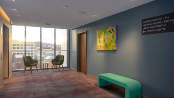 A modern hotel lobby with a large window, two green armchairs, a small round table, and a teal bench. A colorful abstract painting adorns the wall, and directional signage indicates guest rooms and facilities.