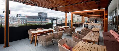 An outdoor terrace with wooden tables and wicker chairs, a bar area, and a city view.