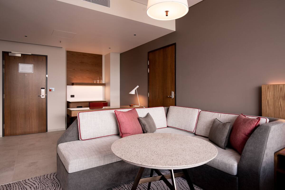 Une chambre d’hôtel moderne avec un canapé d’angle gris, une table ronde et un meuble en bois.
