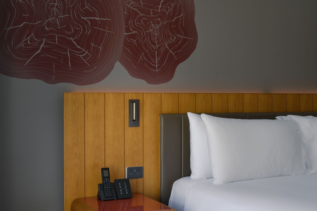 Habitación de hotel con cabecera de madera, cama con ropa blanca, mesita de noche con teléfono y control remoto, y lámpara fijada a la pared.