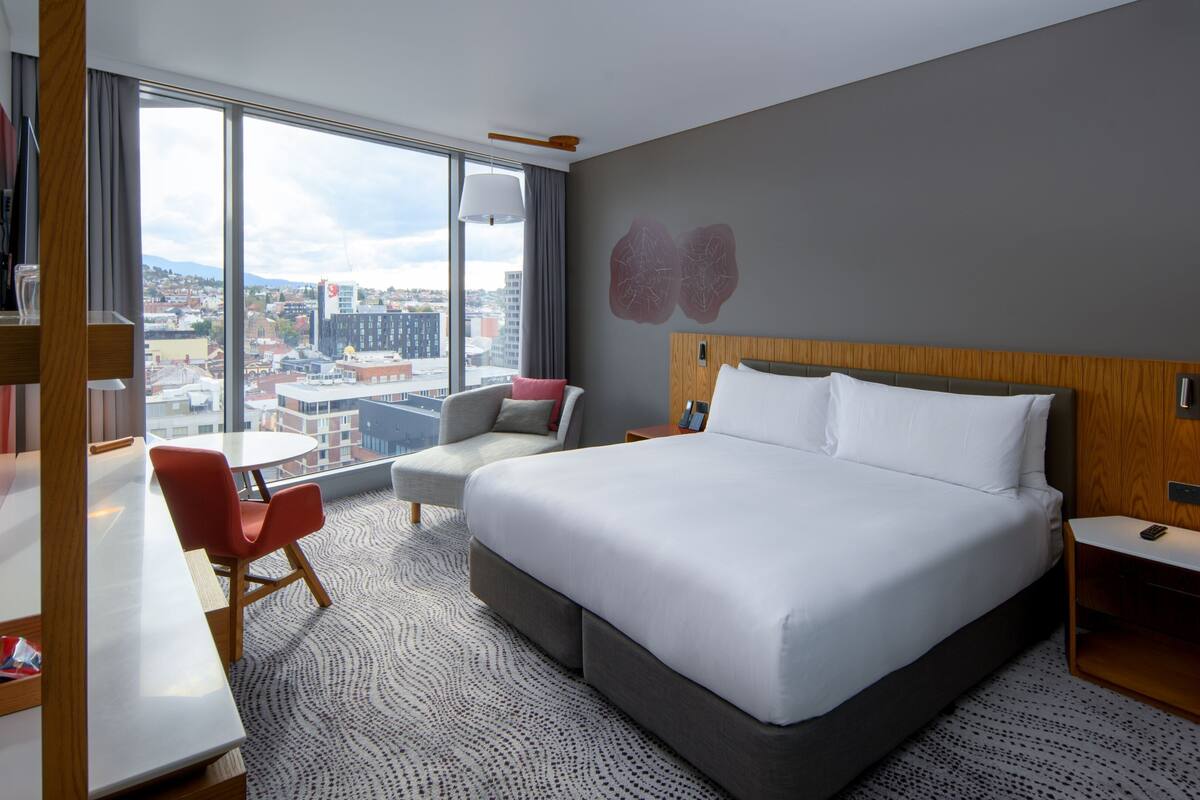Une chambre d’hôtel avec un grand lit, un bureau, une chaise et une vue sur la ville grâce à de grandes fenêtres.