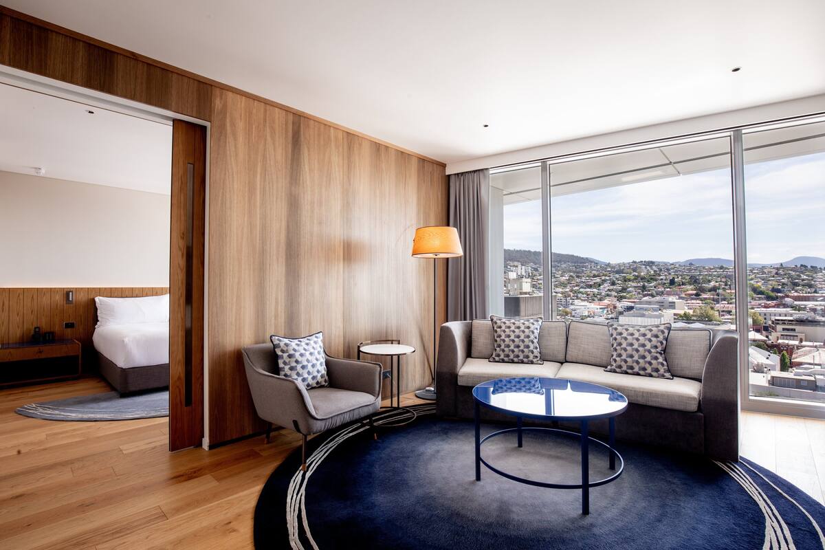 Une chambre d’hôtel moderne avec une grande fenêtre, un canapé, un fauteuil, une petite table et un lit.