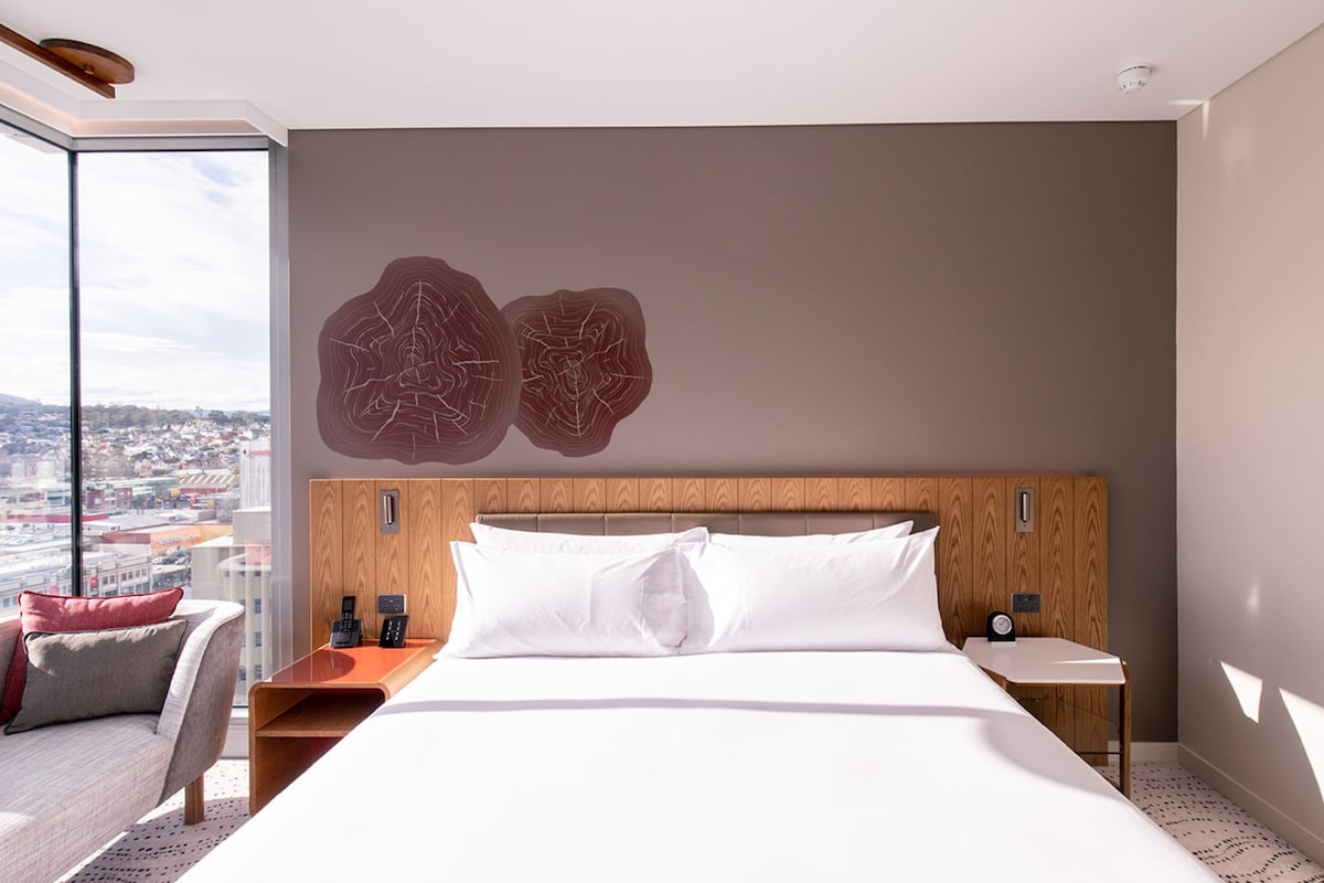 Habitación de hotel moderna con una cama grande, cabecera de madera, vistas a la ciudad y una pared de vidrio.