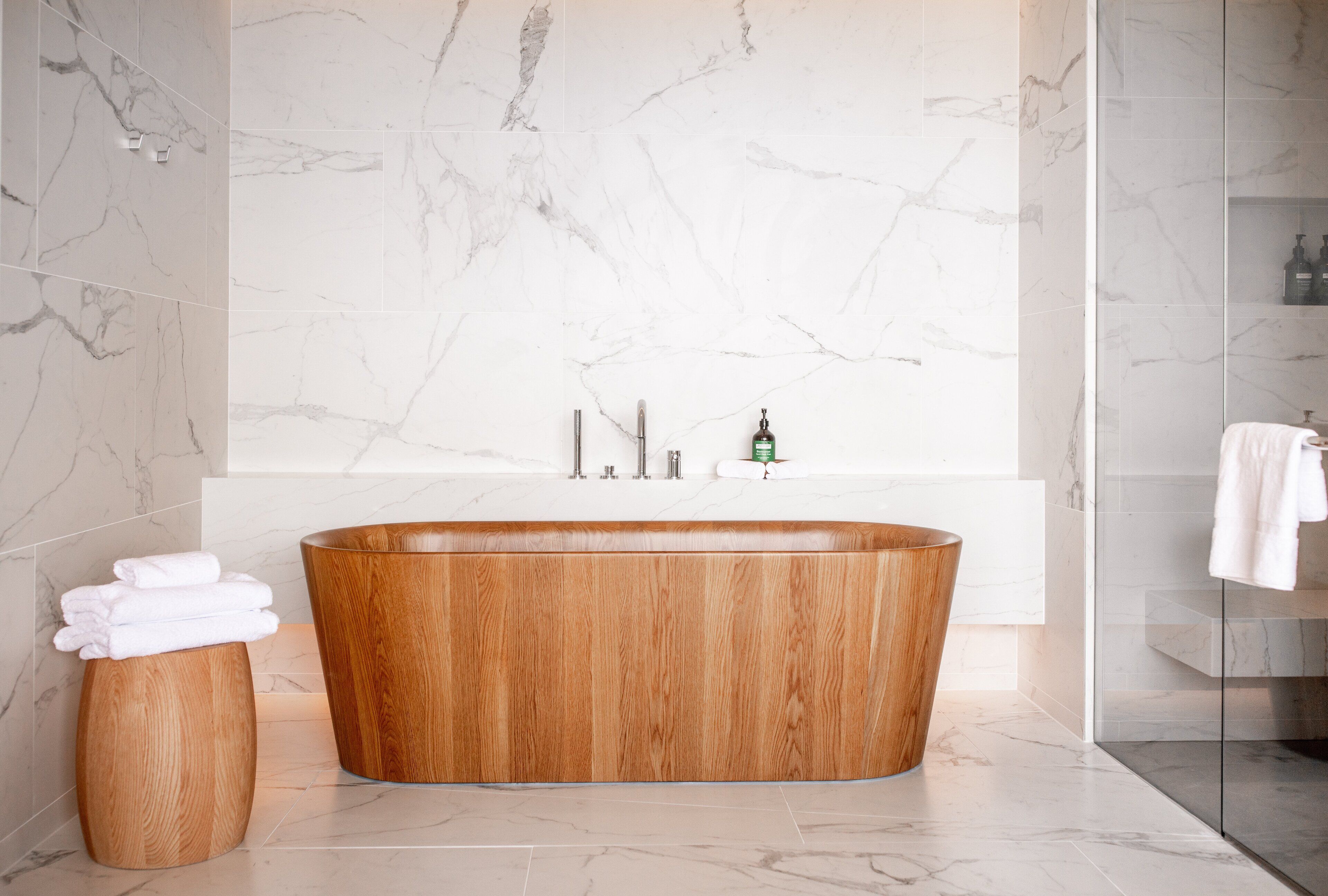 A modern bathroom with a freestanding bathtub, marble walls, and a wooden stool with towels.